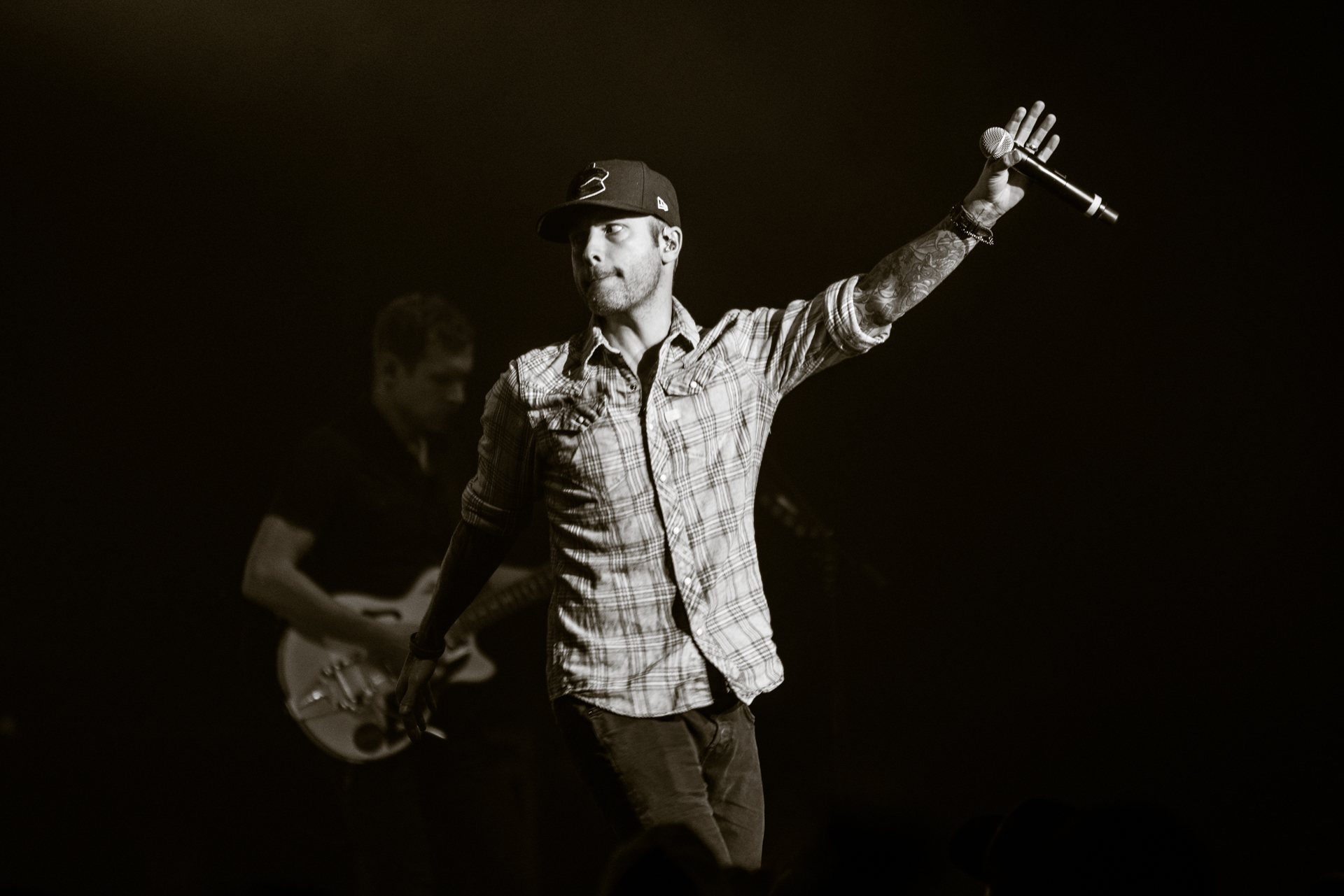 Dallas Smith commands the stage, holding the microphone high in his left hand as he scans the crowd with an electric energy. In the background, a man passionately plays the electric guitar, adding to the intensity of the moment. The black and white photo captures the dynamic and emotive essence of the performance.