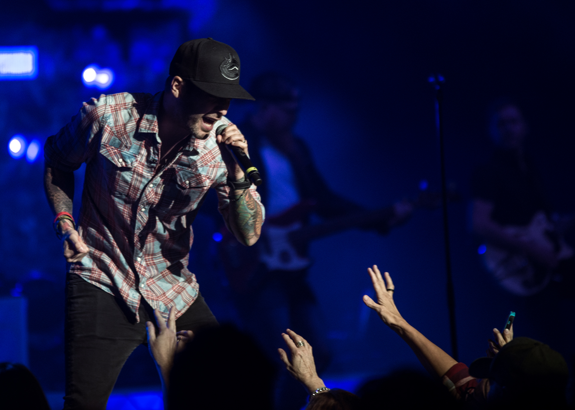Dallas Smith, clad in a red and green plaid shirt and a baseball cap, takes the stage, singing into a microphone. The energy is palpable as the audience reaches out towards him, creating a dynamic connection between the artist and the crowd.
