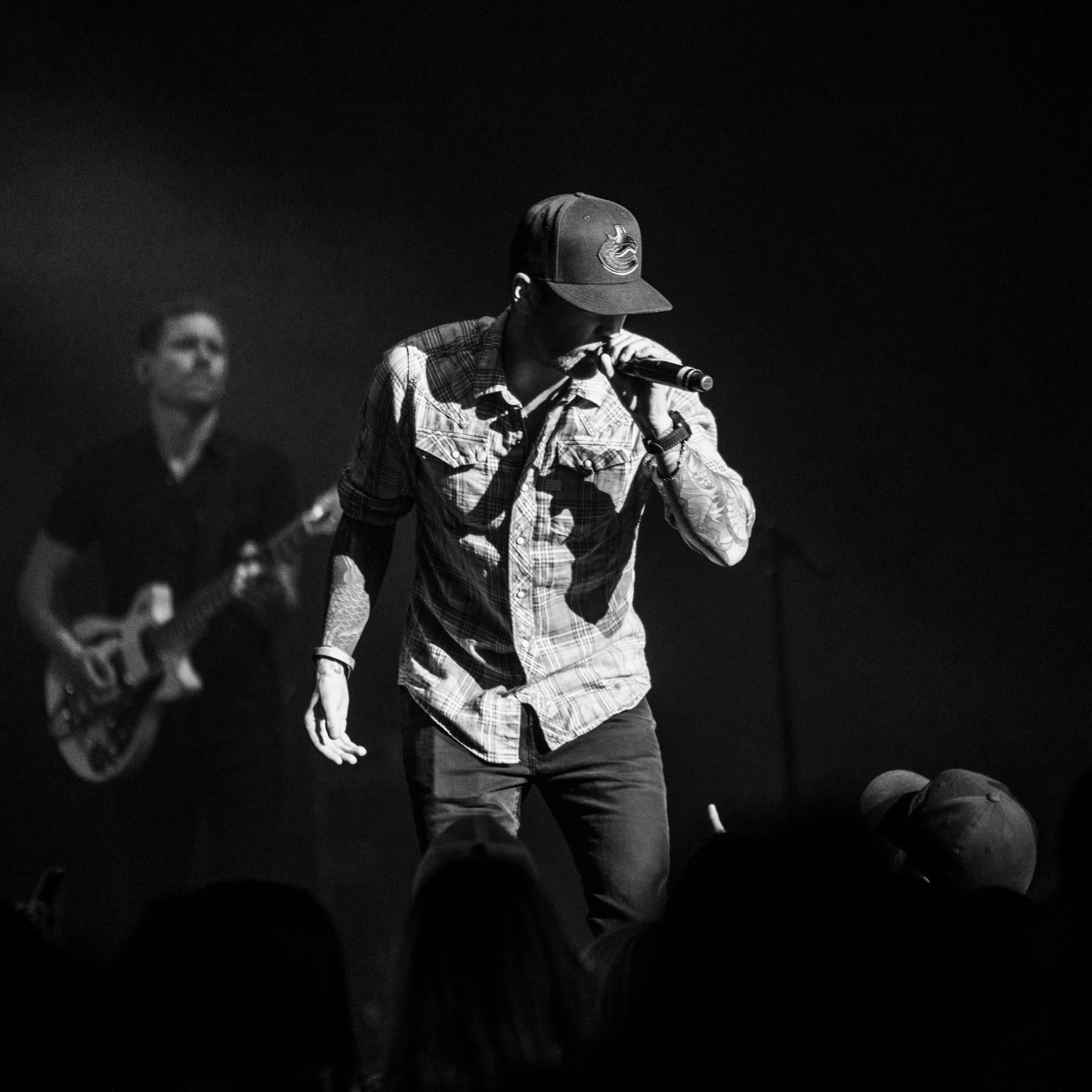 Dallas Smith takes center stage, singing into a microphone, while in the background, a man passionately plays an electric guitar. The black and white photo captures the essence of the musical performance, emphasizing the timeless and emotive nature of the scene.