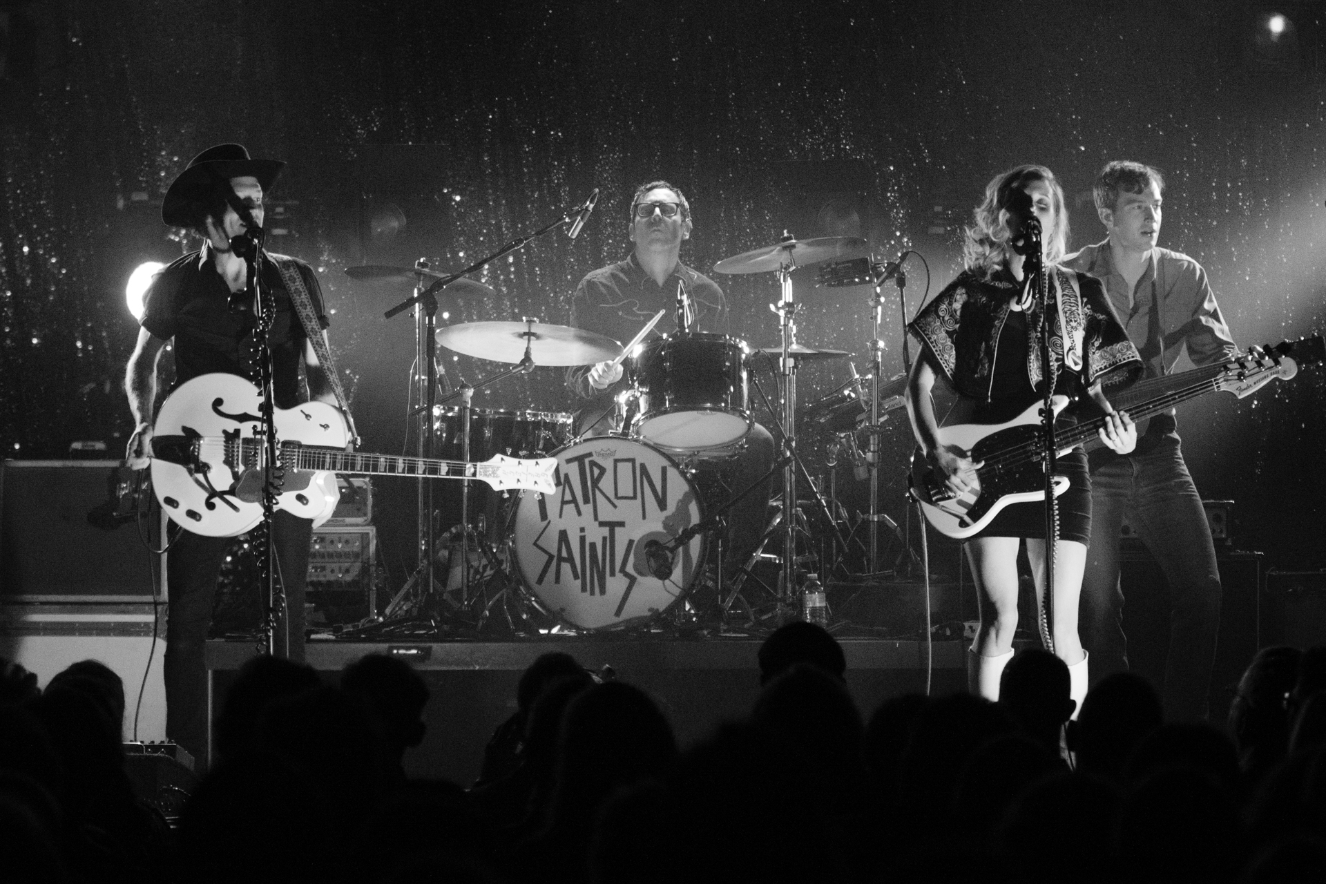 Whitehorse band members create a timeless tableau on stage in a black and white photo. Luke Doucet on the left strums an acoustic guitar, Melissa McClelland on the right wields an electric guitar, while a drummer holds the center stage. In the background, another band member plays a guitar, capturing the collaborative and harmonious essence of their musical performance.
