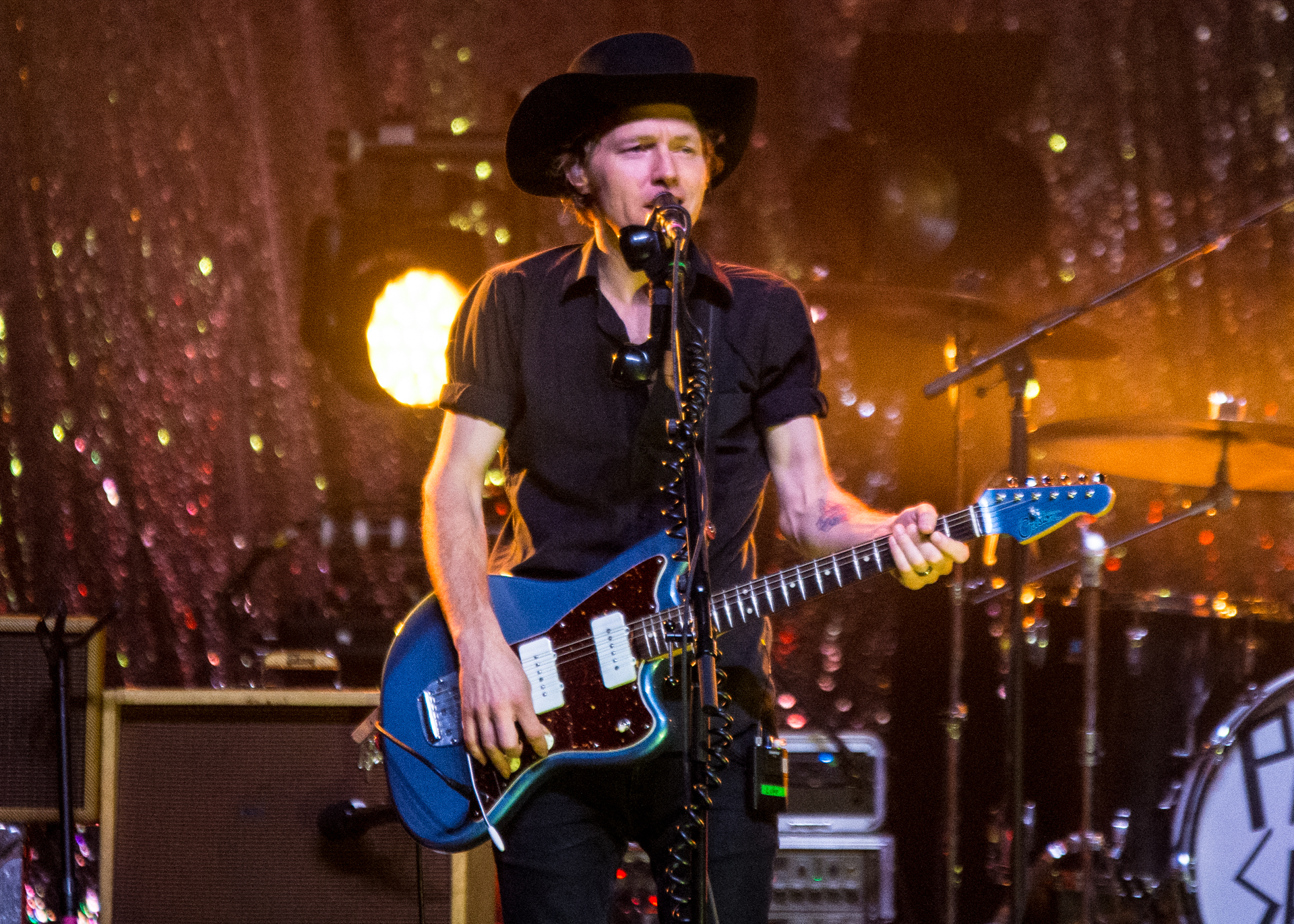Luke Doucet, sporting a cowboy hat, commands the stage, holding his blue electric guitar and singing into the microphone. The spotlight illuminates him against a sparkly backdrop, creating a visually striking and dynamic presence that captures the essence of his performance.