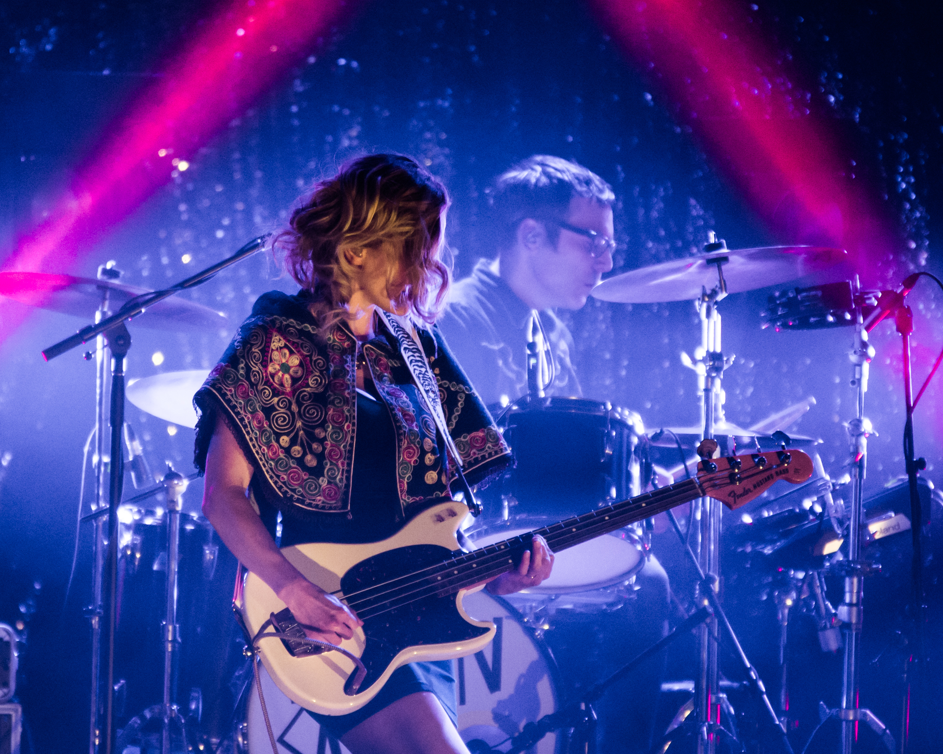 Melissa McClelland takes the stage in vibrant style, adorned in a brightly colored patterned shawl, playing a white electric guitar. The drummer provides a rhythmic backdrop, while the blue-lit stage is framed by two neon pink light rays, creating a visually dynamic and energetic scene.