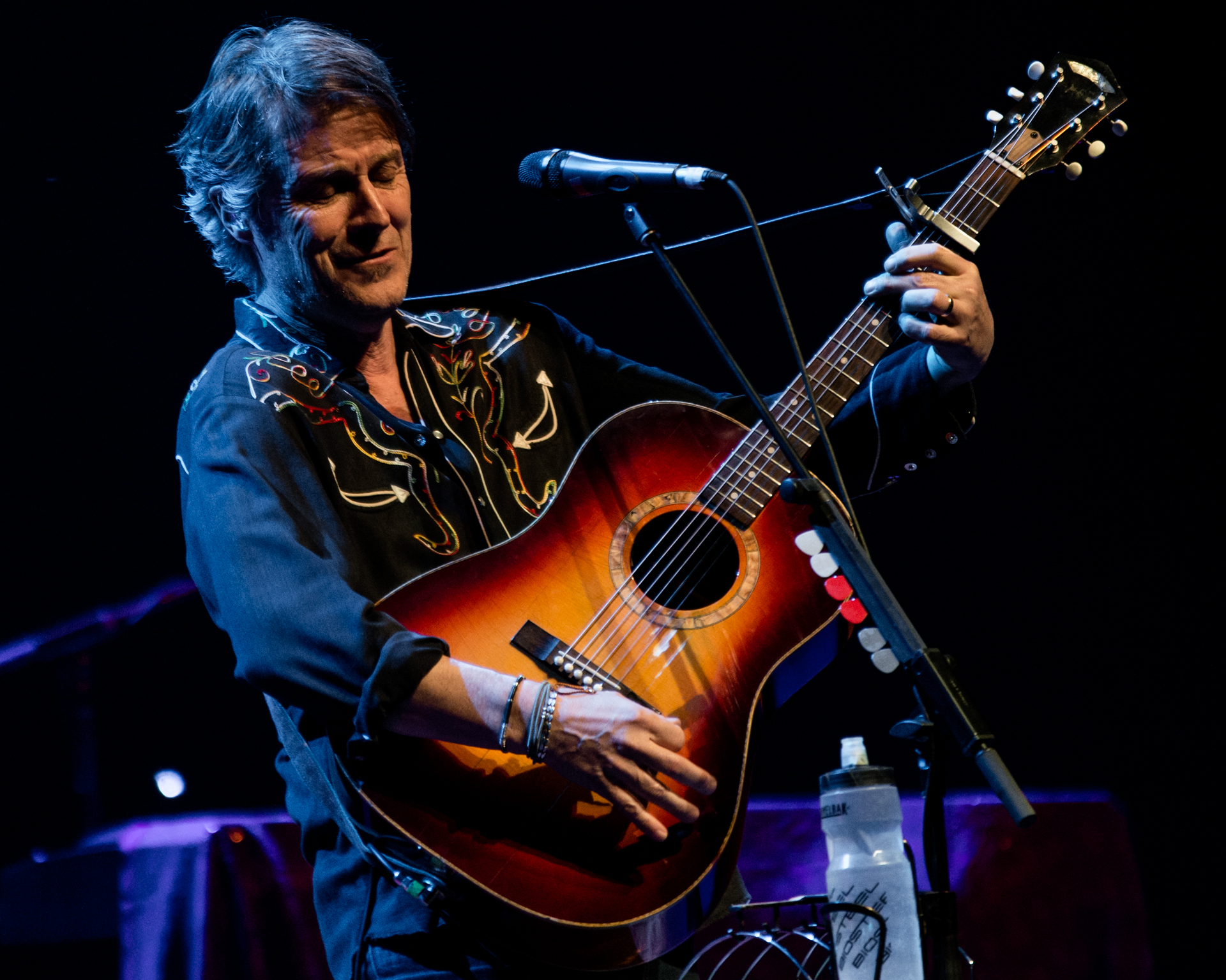 Jim Cuddy commands the stage in a black collared shirt with unique stitching, skillfully playing an acoustic guitar. Notably, his signature water bottle is attached to the microphone stand, a distinctive touch adding a personal flair to the performance. 
