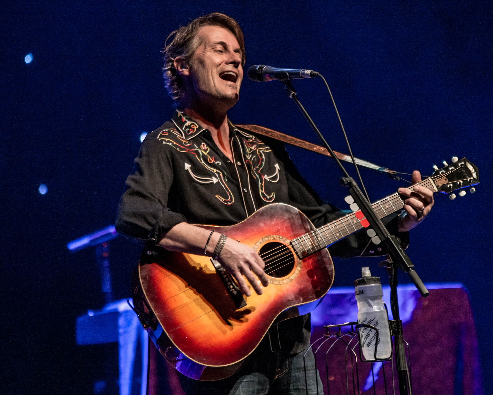 Jim Cuddy commands the stage in a black collared shirt with unique stitching, singing into the microphone and skillfully playing an acoustic guitar. Notably, his signature water bottle is attached to the microphone stand, a distinctive touch adding a personal flair to the performance. 