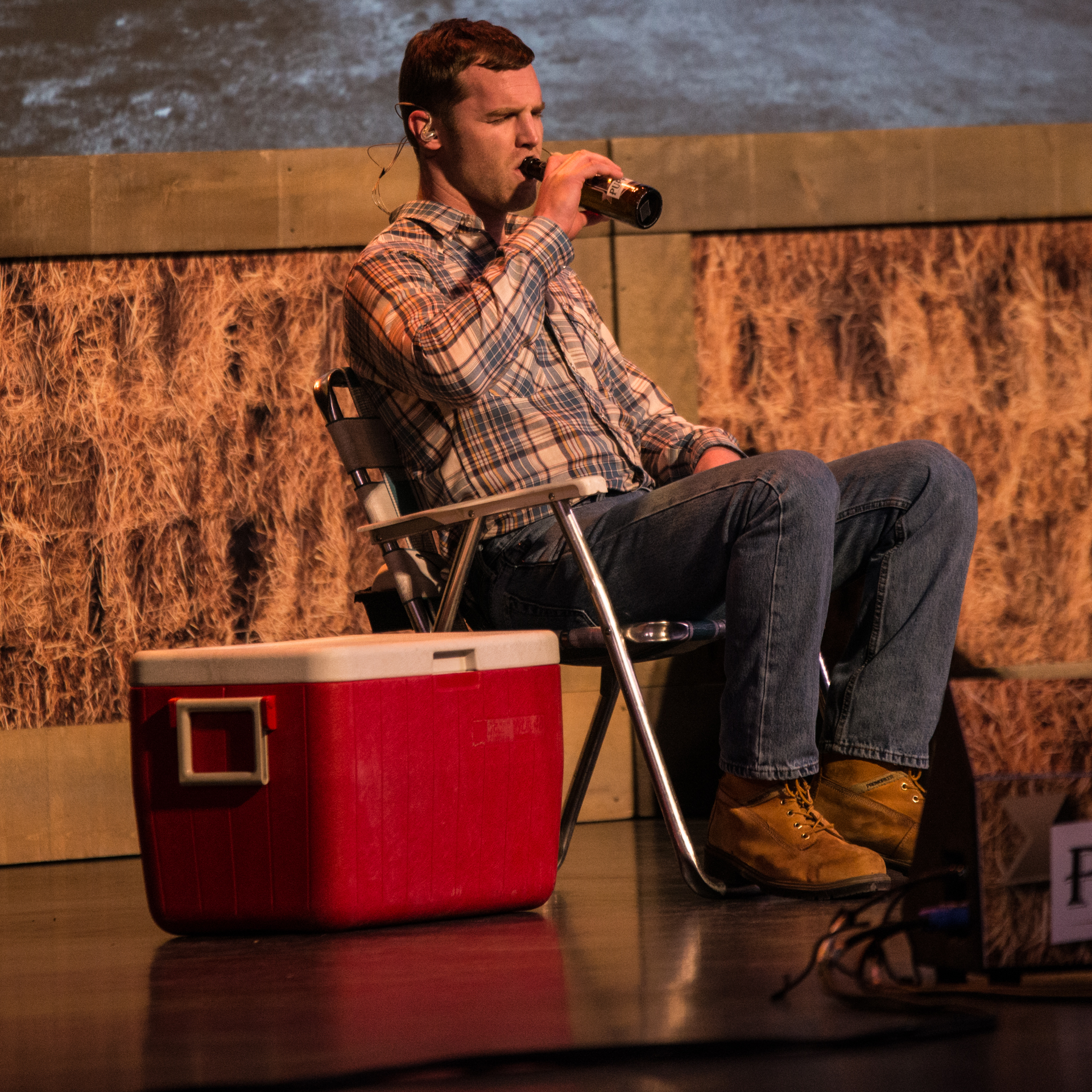 Jared Keeso sits comfortably on stage, donned in a blue and white plaid shirt and blue jeans, taking a sip of Puppers beer. Beside him, a red cooler adds a touch of character. The backdrop of hay completes the rustic and inviting atmosphere, setting the stage for a unique and enjoyable performance.