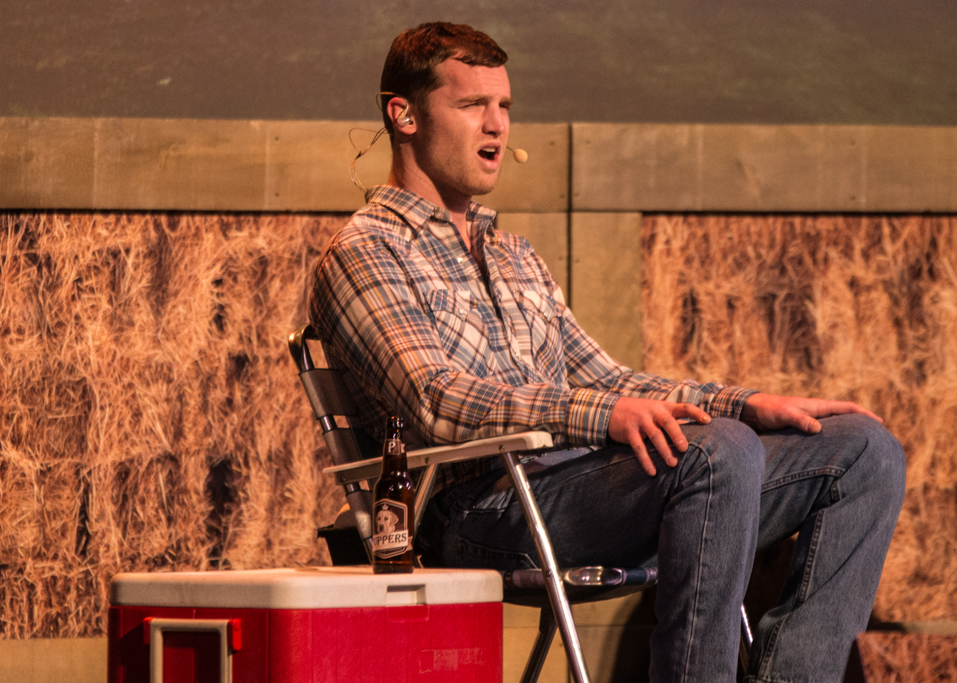 Jared Keeso sits comfortably on stage, donned in a blue and white plaid shirt and blue jeans, engaging with the audience through a headworn microphone. Beside him, a red cooler with a Puppers beer atop adds a touch of character. The backdrop of hay completes the rustic and inviting atmosphere, setting the stage for a unique and enjoyable performance.