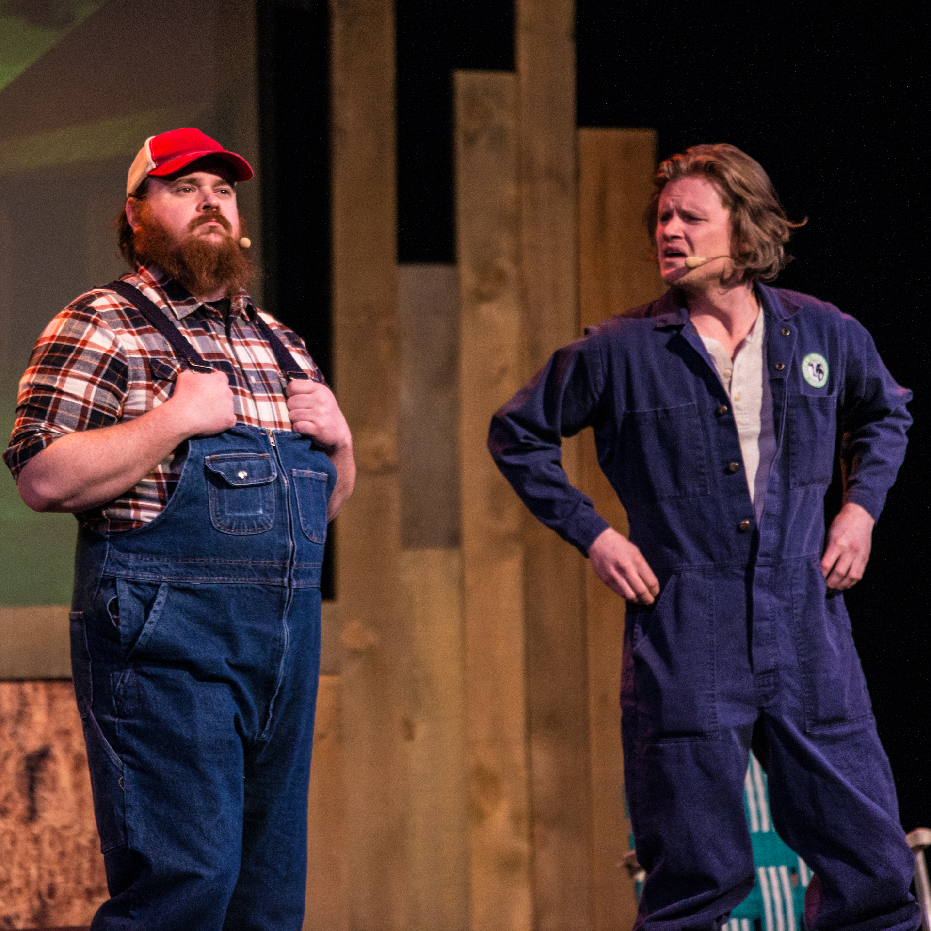 Nathan Dales and K. Trevor Wilson share a comically confused moment on stage. Nathan, hands on hips, addresses K. Trevor with a quizzical expression, while K. Trevor, holding onto the straps of his overalls, gazes off into space. Their playful interaction adds a touch of humor to the performance, creating an entertaining and engaging scene.