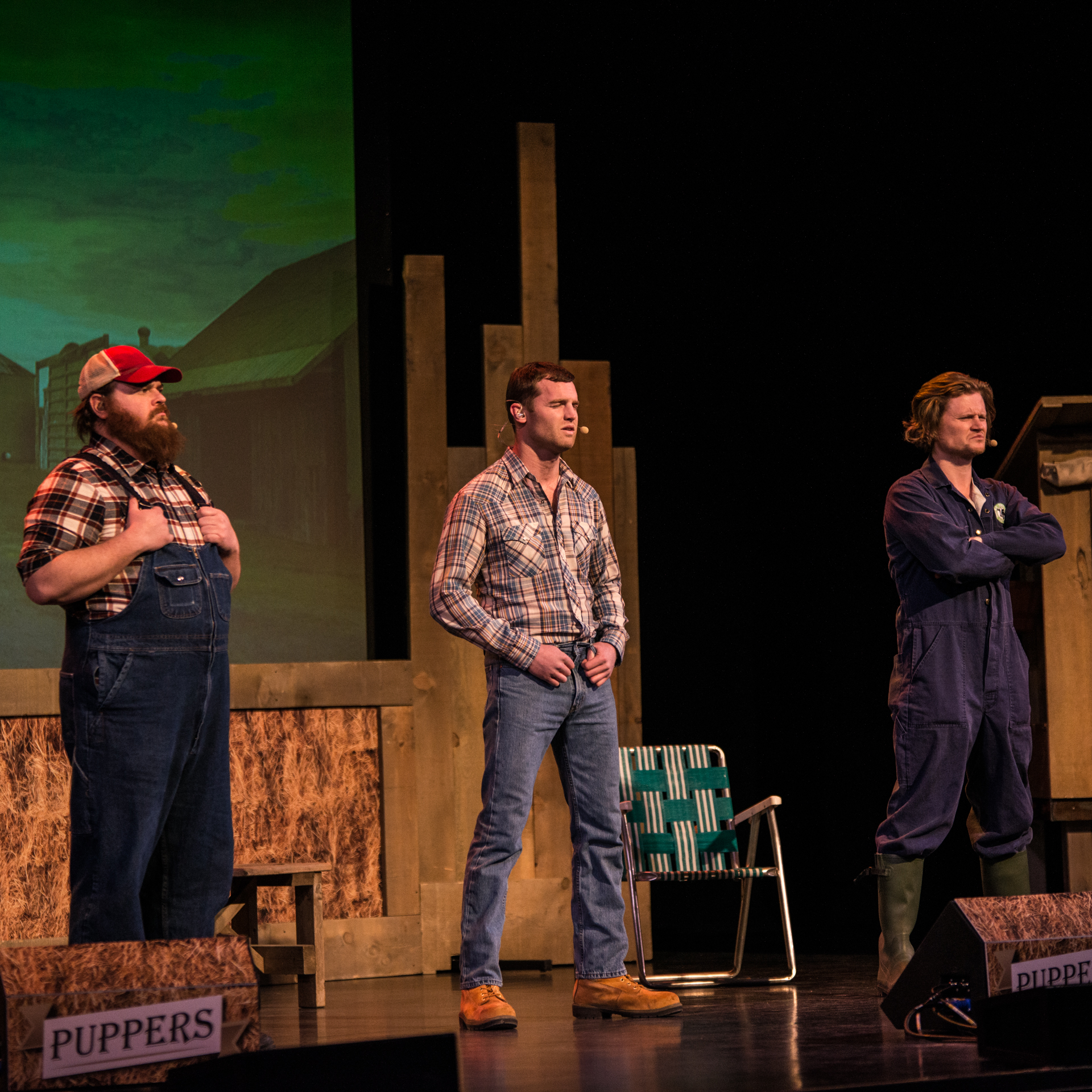 Jared Keeso takes center stage, gripping the front loops of his blue jeans, flanked by Nathan Dales on the right, arms folded, and K. Trevor Wilson on the left, clutching the straps of his overalls. The backdrop features hay, a green folding chair, and the foreground is adorned with a Puppers sign, creating a rustic and visually engaging setting for their performance.
