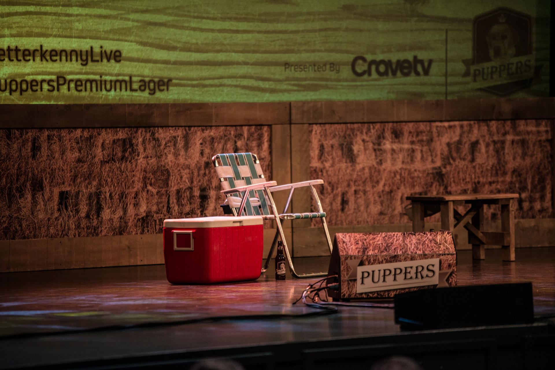 The stage is set with a solitary green folding chair, a red cooler on the left of the chair, a Puppers beer underneath the chair, and a prominent Puppers sign in the foreground. In the background, hay contributes to the rustic atmosphere, alongside a promotional sign advertising Letterkenny on Crave TV. The scene is a perfect blend of laid-back charm and entertainment.