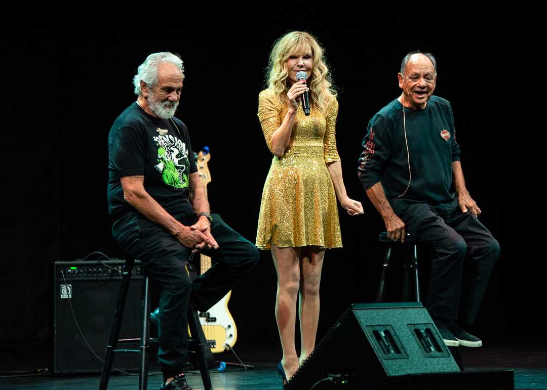Tommy Chong and Cheech Marin sit on stools on stage, flanking Shelby Chong in the middle. Shelby, adorned in a gold, sparkly dress, holds a microphone, addressing the crowd with charm and charisma.