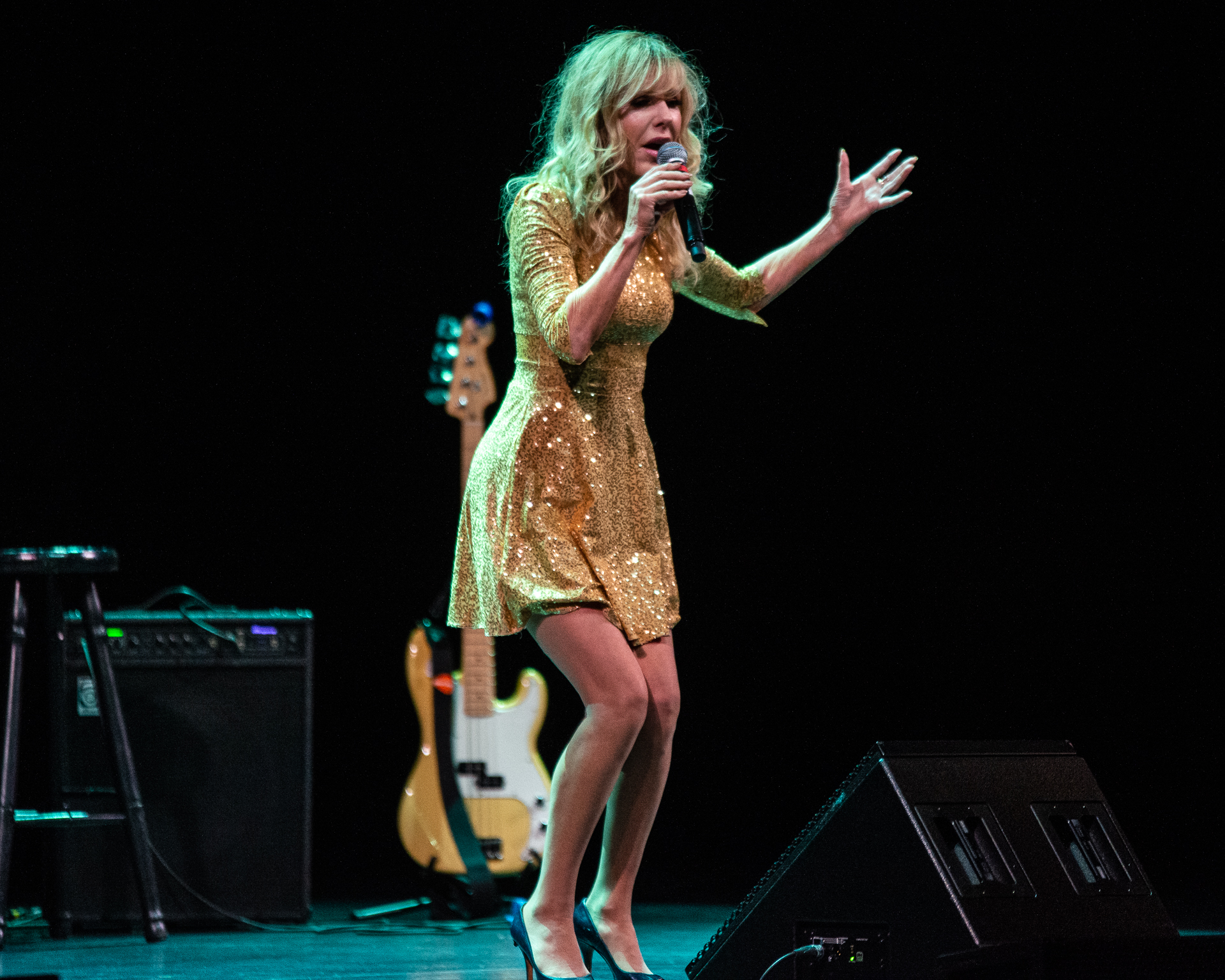 Shelby Chong commands the stage in a dazzling gold, sparkly dress, wielding a microphone with expressive body movements as she addresses the crowd. In the background, a yellow electric guitar adds to the vibrant atmosphere of the performance.
