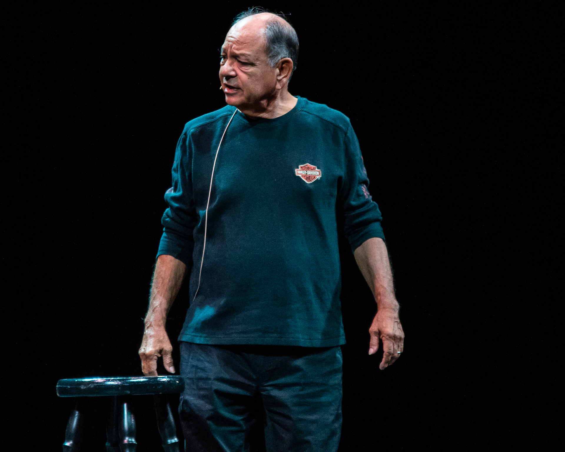 Cheech Marin stands on stage beside a stool, sporting a Harley Davidson long sleeve shirt. His expression is quizzical, adding a touch of curiosity and intrigue to the atmosphere.