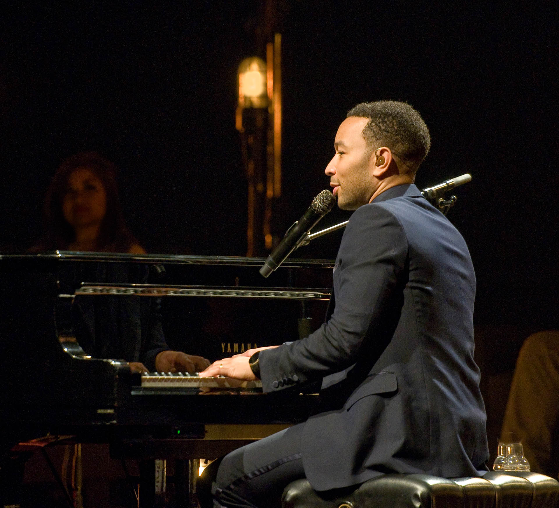 John Legend, elegantly attired in a black suit, sits at a black Yamaha piano, delivering a soulful performance on stage. The soft glow of white light surrounds him, creating a captivating atmosphere, as blissful audience members watch his performance with delight.