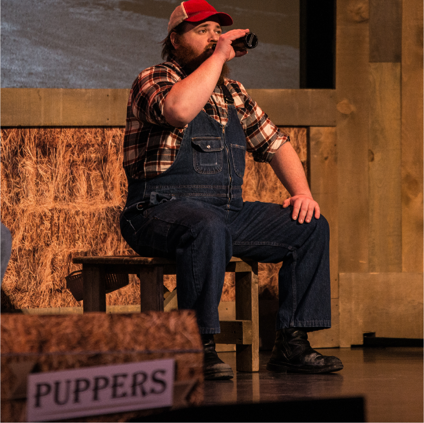 K. Trevor Wilson sits casually on a wooden stool, donned in navy blue overalls, a red and black plaid shirt, and a red baseball cap. With an engaging presence, he takes a sip of Puppers beer, creating a laid-back and authentic atmosphere. The background features hay, and in the foreground, a Puppers sign adds to the rustic charm of the scene.
