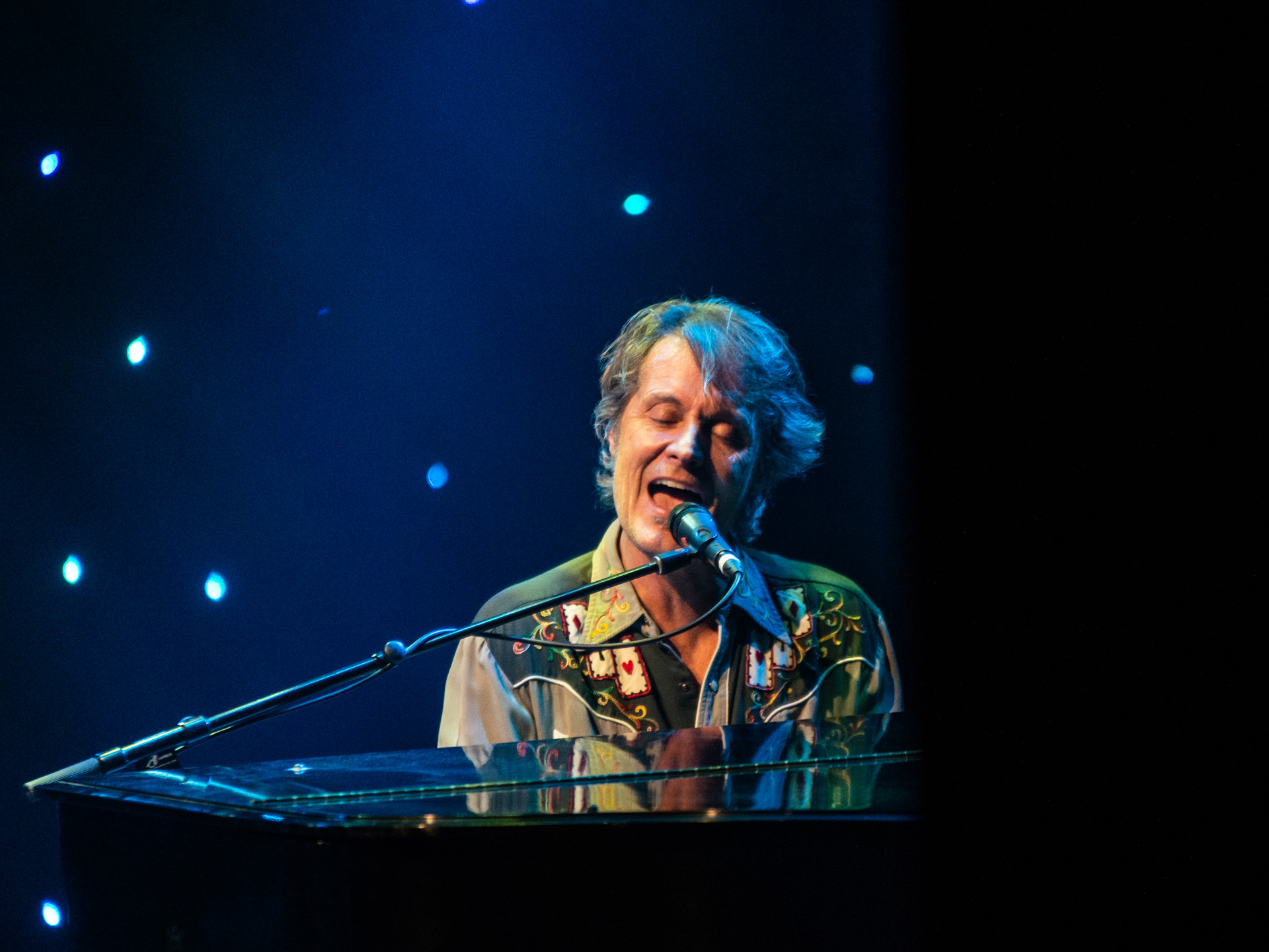 Jim Cuddy captivates the audience as he sings into the microphone while skillfully playing the piano. Behind him, a mesmerizing display of globes of lights adds a touch of magic to the stage, creating a visually enchanting atmosphere for his performance.