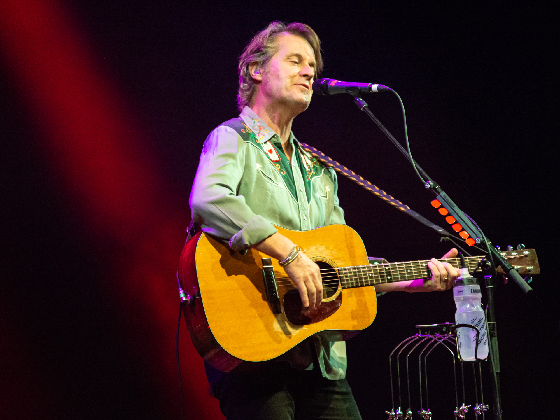 Jim Cuddy commands the stage in a vibrant green collared shirt, singing into the microphone and skillfully playing an acoustic guitar. A red spotlight illuminates him from the back, casting a dramatic aura. Notably, his signature water bottle is attached to the microphone stand, a distinctive touch adding a personal flair to the performance. 