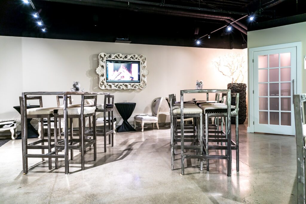 In the lounge, two sets of stylish tables with chic chairs in chrome trim take center stage, creating an inviting setting. A TV framed with a decorative frame adorns the wall in the background, adding a modern touch. To the right, a glass door brings in additional light, while track lights in the ceiling brightly illuminate the space, enhancing its contemporary and welcoming atmosphere.