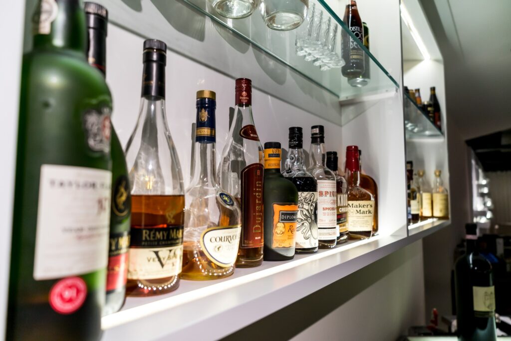 In the lounge bar, a row of upscale alcohol bottles adorns a shelf to the left, accompanied by a glass shelf above it featuring neatly arranged glassware. The scene is brightly lit from above, accentuating the sophistication of the space and highlighting the premium selection of beverages.