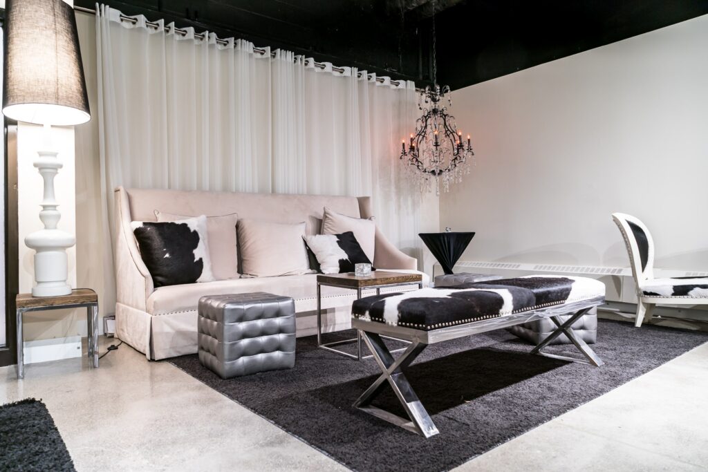 The lounge features a sophisticated ambiance with a white couch adorned with black and white pillows, a silver ottoman, and a wooden table, complemented by a black and white bench seat, all arranged on a sleek black carpet. A curtain wall serves as a stylish backdrop. To the left, a large lamp adds to the contemporary design, while a chandelier hangs from the ceiling on the right, contributing to the bright and inviting atmosphere of the space.