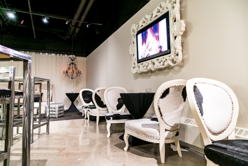 The lounge exudes a modern and elegant ambiance with bright white walls and beige floors. White chairs with black spots and a table covered in a black tablecloth are arranged on the right, creating a stylish seating area. A TV framed with a decorative frame adorns the right wall, adding a touch of contemporary design. In the far back, a chandelier hangs from the ceiling, softly emitting a warm yellow light, contributing to the overall chic atmosphere. The space is brightly illuminated with spotlights, completing the sophisticated and inviting setting.