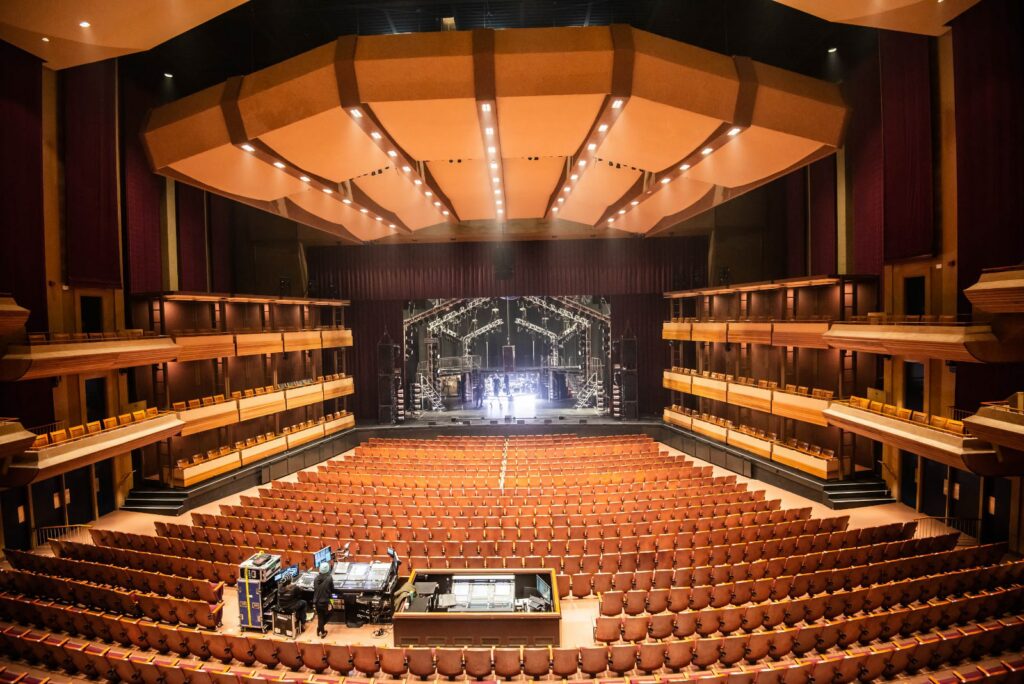 The theatre reveals an expansive centered view from the back, showcasing 2000 empty burnt orange seats with wood trim filling the auditorium. The empty stage in the background awaits performances. The space is brightly lit by numerous pot lights in the ceiling. A centrally located sound booth adds to the technical setup. Boxed seating along the sides of the theatre, adorned with wood trim, contributes to the overall aesthetic of this spacious and well-appointed venue.