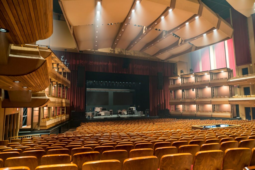 The theatre reveals an expansive view from the back, showcasing 2000 empty burnt orange seats with wood trim filling the auditorium. The empty stage in the background awaits performances. The space is brightly lit by numerous pot lights in the ceiling. A sound booth to the right adds to the technical setup. Boxed seating along the sides of the theatre, adorned with wood trim, contributes to the overall aesthetic of this spacious and well-appointed venue.