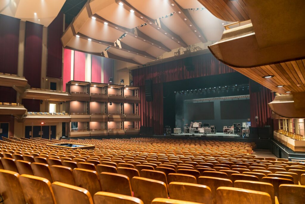 The theatre reveals an expansive view from the back right, showcasing 2000 empty burnt orange seats with wood trim filling the auditorium. The empty stage in the background awaits performances. The space is brightly lit by numerous pot lights in the ceiling. A sound booth on the left adds to the technical setup. Boxed seating along the sides of the theatre, adorned with wood trim, contributes to the overall aesthetic of this spacious and well-appointed venue.