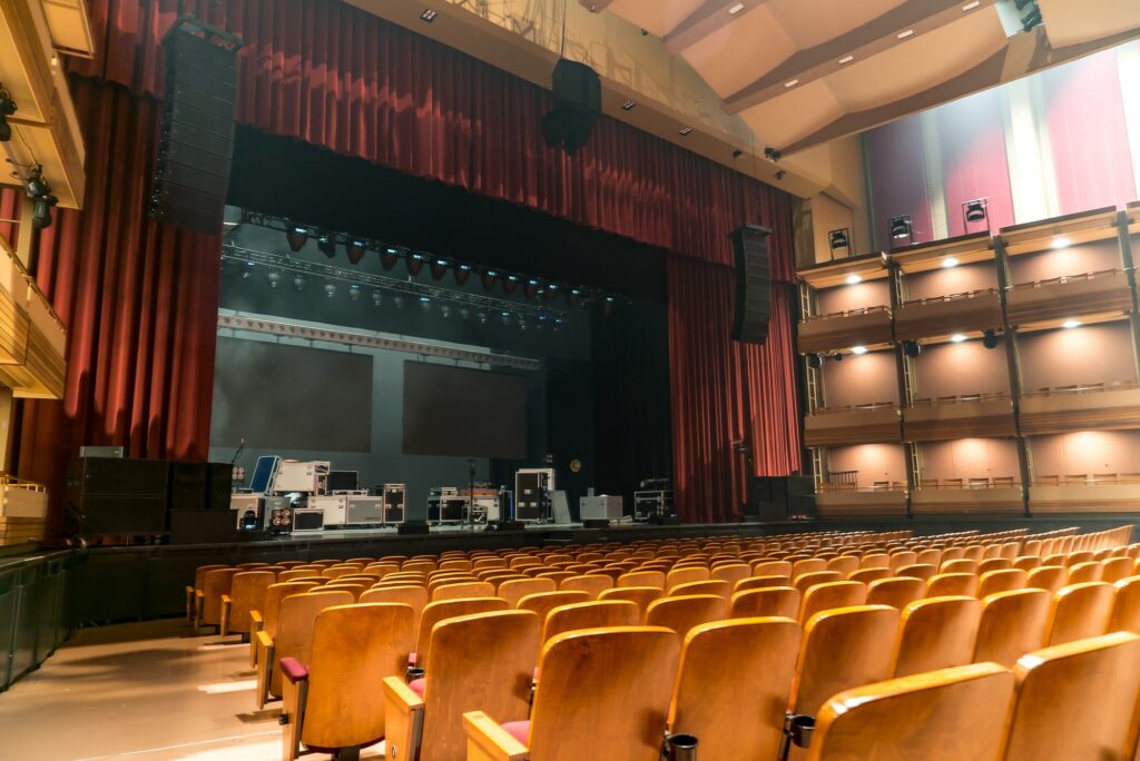 The theatre reveals an expansive view from the back left, showcasing 2000 empty burnt orange seats with wood trim filling the auditorium. The empty stage in the background awaits performances. The space is brightly lit by numerous pot lights in the ceiling. Boxed seating along the sides of the theatre, adorned with wood trim, contributes to the overall aesthetic of this spacious and well-appointed venue.