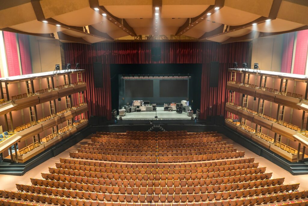 The theatre reveals an expansive view from the back, showcasing 2000 empty burnt orange seats with wood trim filling the auditorium. The empty stage in the background awaits performances. The space is brightly lit by numerous pot lights in the ceiling. Boxed seating along the sides of the theatre, adorned with wood trim, contributes to the overall aesthetic of this spacious and well-appointed venue.
