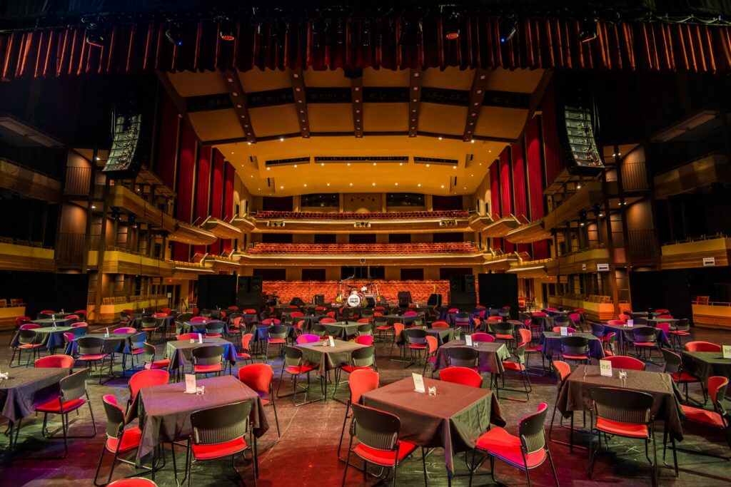 The stage is set with elegance, featuring tables adorned in black cloths and complemented by red chairs, each table meticulously set with neatly placed menus for a sophisticated event. In the background, musical instruments stand ready, adding a touch of anticipation, while the empty theatre seats create a unique and inviting atmosphere for the upcoming performance.