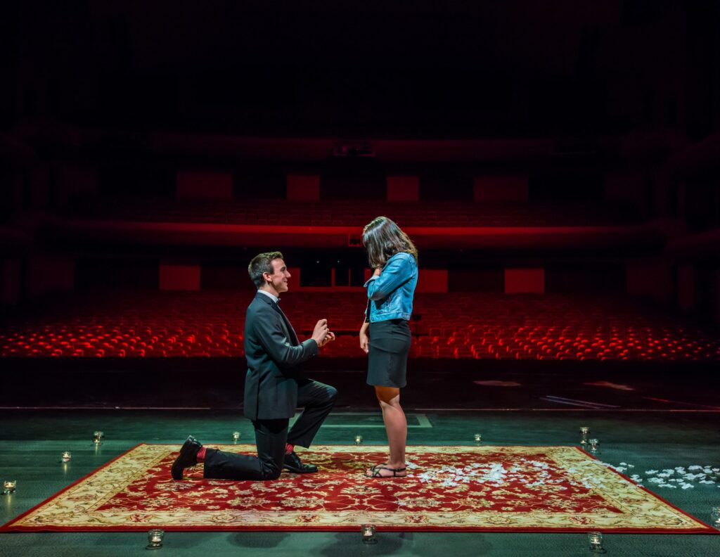 On a stage adorned with a carpet covered in white petals, a man kneels down on one knee, proposing to his girlfriend. The romantic scene unfolds against the backdrop of empty seats, creating an intimate and heartfelt moment in a seemingly private setting.