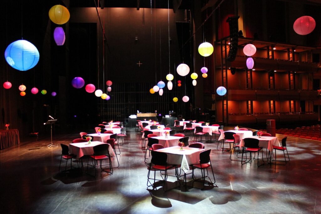 The stage is transformed into a vibrant setting with tables and chairs arranged, adorned with colourful fabric balloons hanging from the ceiling. Splashes of light illuminate each table, creating a lively and festive atmosphere, inviting guests to enjoy a dynamic and visually appealing event.