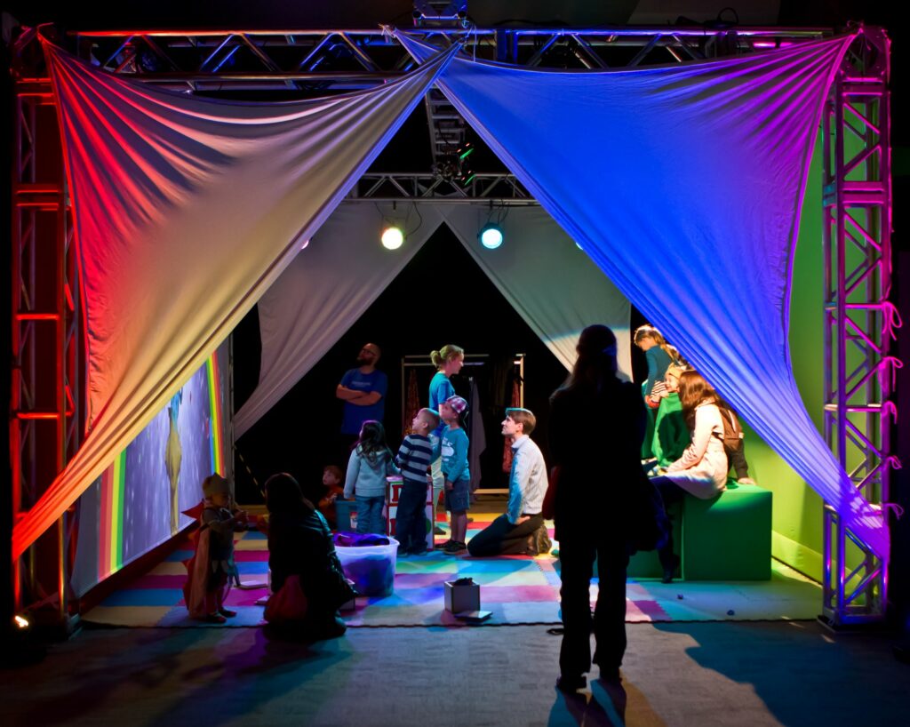 A diverse group of people, including children, stand and sit inside white assembled fabrics, illuminated by multicolored lights. The vibrant play of lights against the white fabric creates a visually captivating and inclusive scene, suggesting a dynamic and immersive environment.